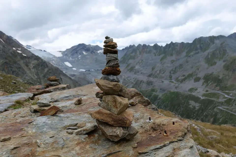 A stone cairn on a rock ledge with a glacier and mountain road behind