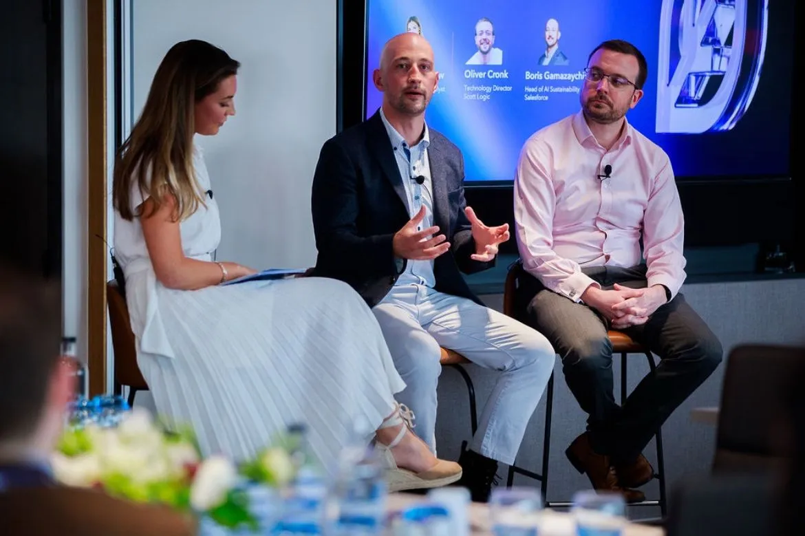 The moderator and two panellists on stools in front of a screen showing the speakers' names, including Oliver Cronk, Technology Director, Scott Logic, and Boris Gamazaychikov, Head of AI Sustainability, Salesforce