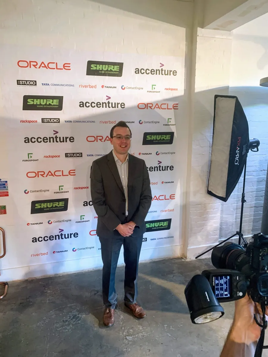 Ollie standing in front of a sponsor backdrop with The Studio and Tanium logos among others, facing a photographer's camera and a softbox light