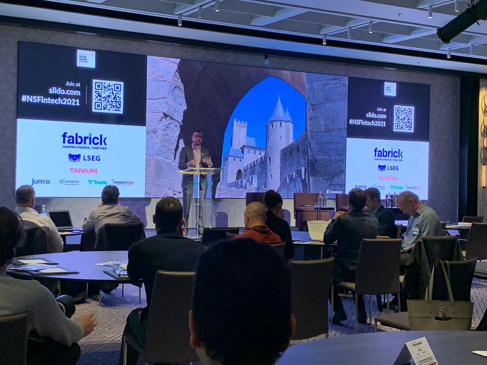 Ollie at a lectern in front of a screen showing a medieval walled castle through a stone archway, flanked by the summit's sponsor logos, including Tanium, and the #NSFintech2021 hashtag