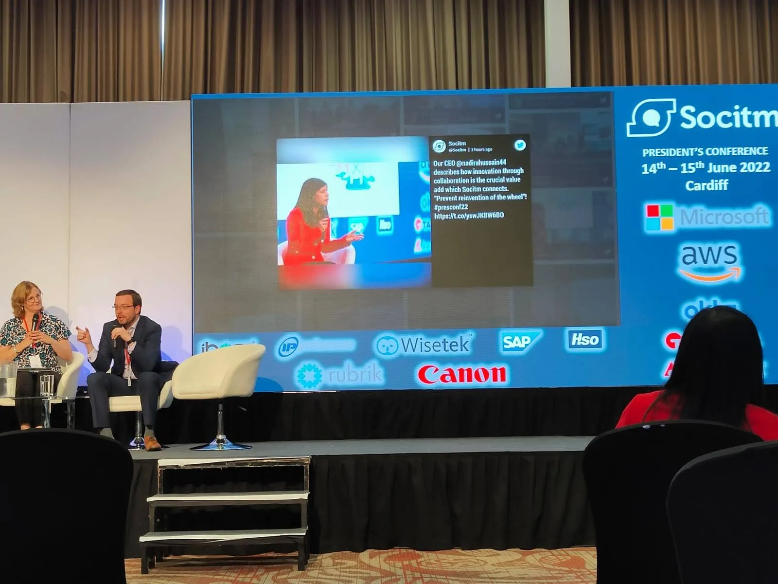 Ollie speaking into a microphone on stage beside his co-presenter, in front of a screen for Socitm's President's Conference, 14 to 15 June 2022, Cardiff