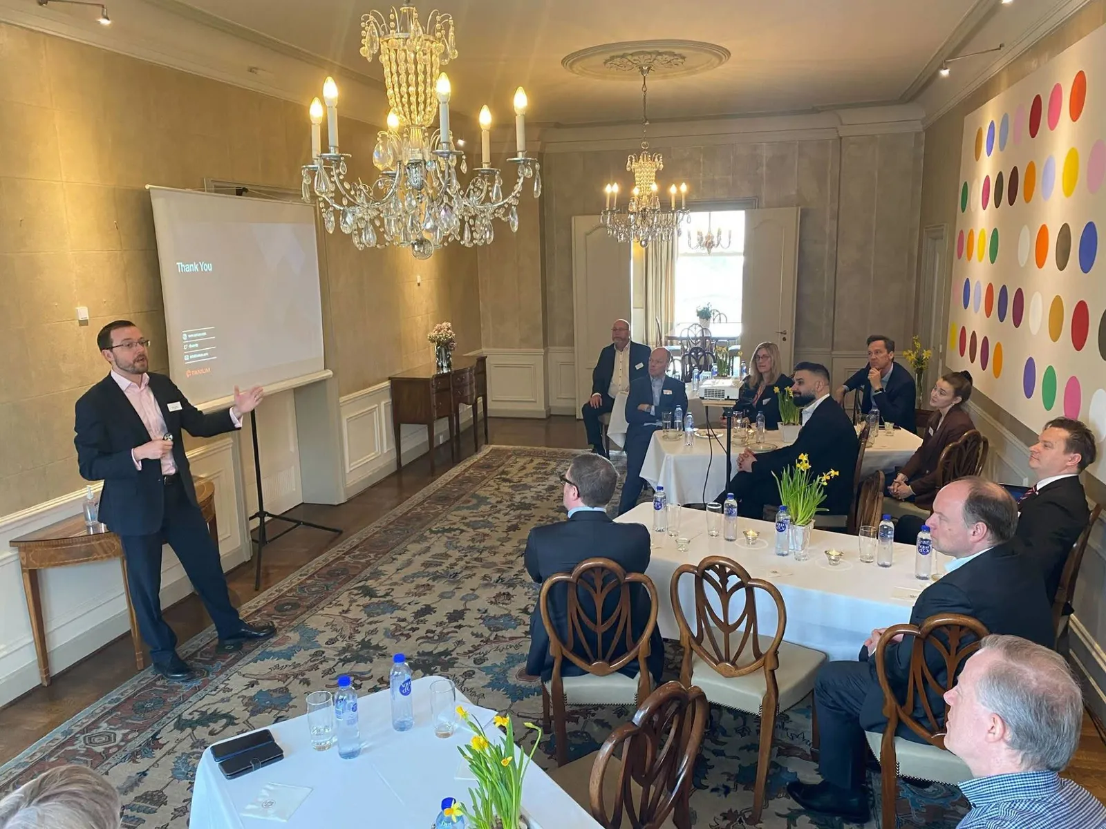 Ollie presenting to guests seated at tables in a room with crystal chandeliers and a wall of coloured spots, with a Tanium thank-you slide on the screen