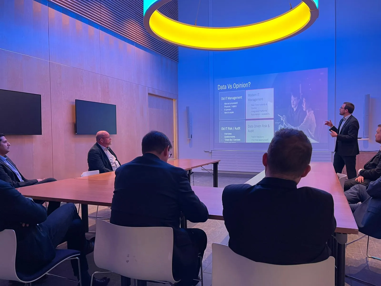 Ollie presenting a slide titled "Data Vs Opinion?" to guests seated around tables, under a large ring-shaped light