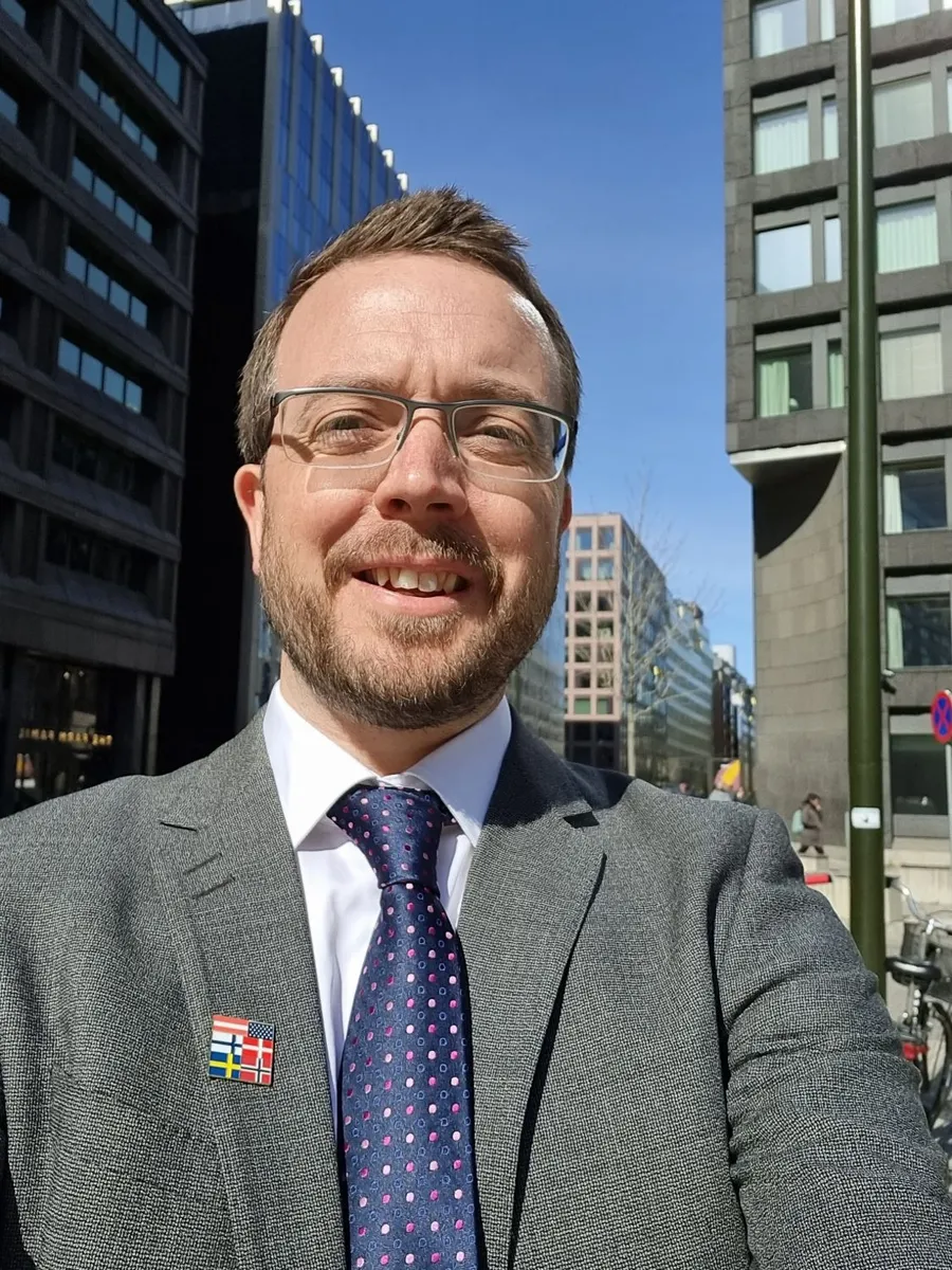 Ollie smiling in a grey suit on a sunny Stockholm street, wearing a lapel pin with the US and Nordic flags