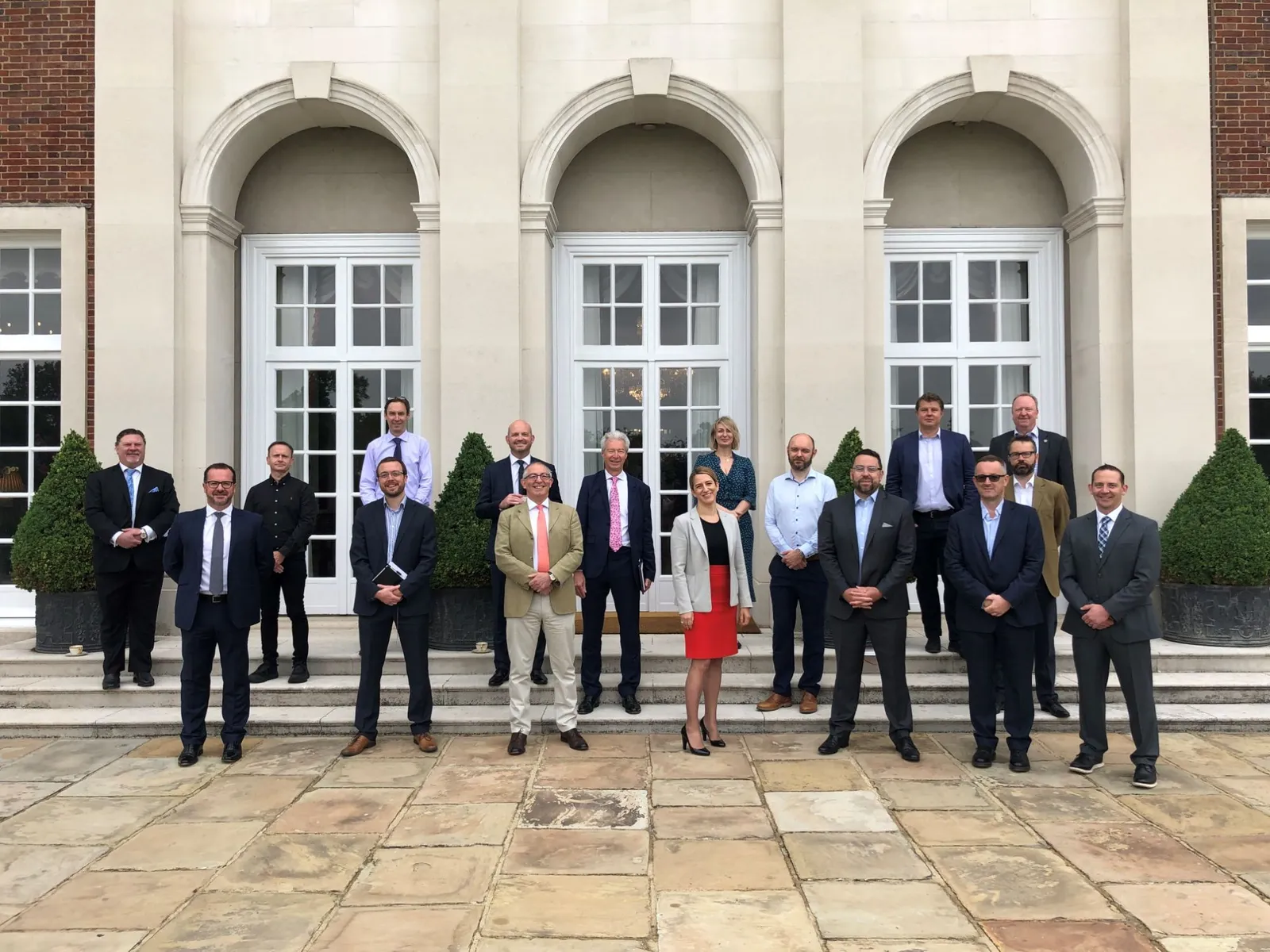 Group photo of about twenty speakers and guests standing on the terrace steps in front of the residence's arched windows